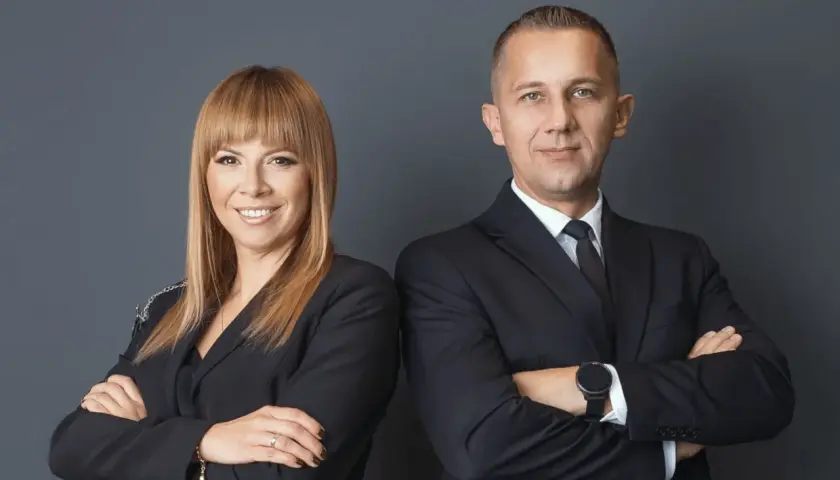Kobieta i mężczyzna w czarnych garniturach, stojący obok siebie ze skrzyżowanymi ramionami.