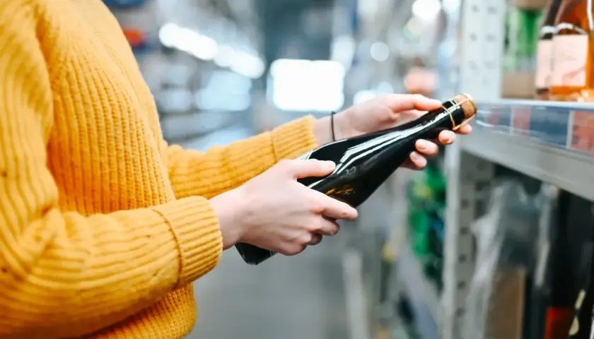 Osoba w żółtym swetrze wybiera butelkę wina w sklepie. Sprawdza ceny alkoholu na Słowacji, porównując dostępne opcje.