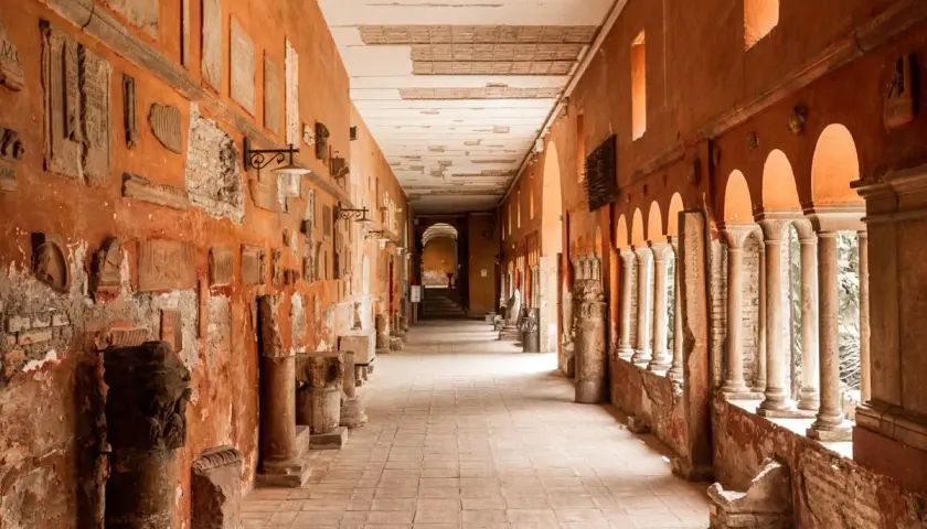 Corridoio con reperti antichi, un'idea di cosa vedere a Trastevere. Colonne, iscrizioni e frammenti decorano le pareti arancioni.