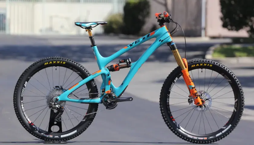A turquoise Yeti SB6c mountain bike with orange accents, featuring Maxxis tires and Fox suspension, ready for any trail.