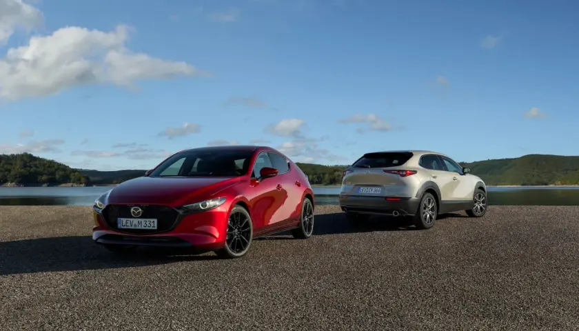 Dwie Mazdy: czerwona hatchback i beżowy SUV, stoją nad jeziorem. Sprawdź mazda cx 30 cena i wybierz swój wymarzony model.
