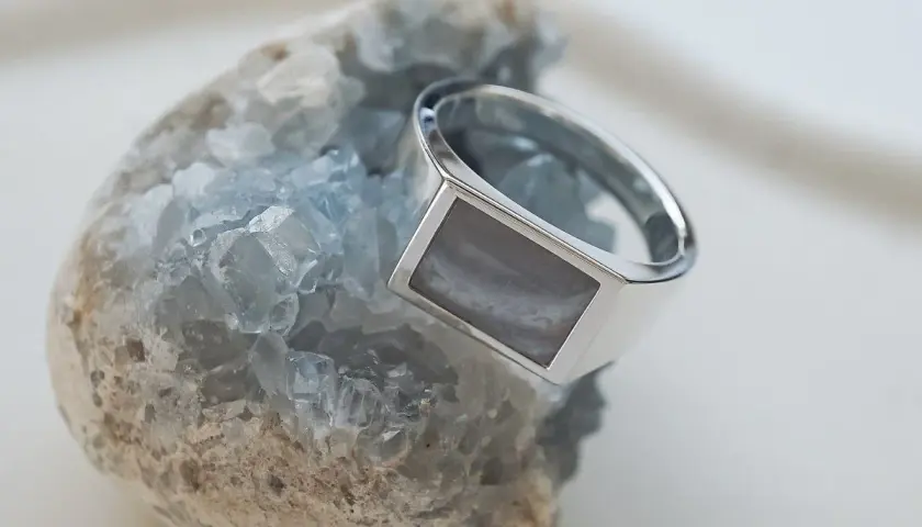 Ein silberner Ring mit einem rechteckigen, grau-weißen Schmuckstein ruht auf einem blauen Kristall.