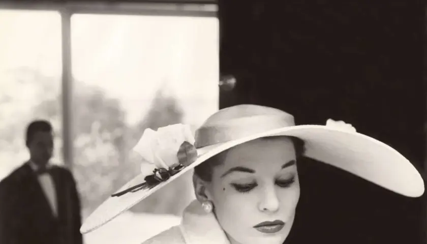 Mujer con sombrero de ala ancha y rosa, estilo Helmut Newton. Un hombre en traje con pajarita al fondo.