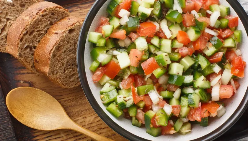 Ensalada pipirrana fresca y colorida, con tomate, pepino, pimiento y cebolla, servida junto a rebanadas de pan integral y una cuchara de madera.