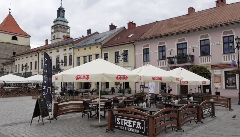 Letni ogródek kawiarniany na rynku w Żywcu. Parasole z logo Żywiec, stoliki i ławki. W tle kamienice i wieża ratuszowa.
