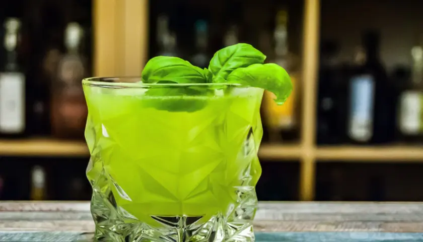 Orzeźwiający drink z likierem miętowym, ozdobiony listkami bazylii, w szklance o geometrycznym kształcie.