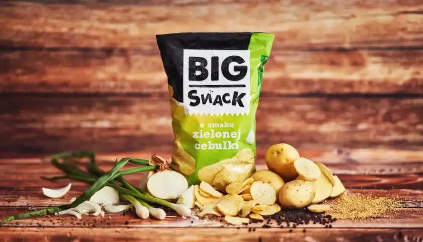 Reklamowa fotografia chipsów Big Snack o smaku zielonej cebulki, z ziemniakami i cebulką na drewnianym tle.