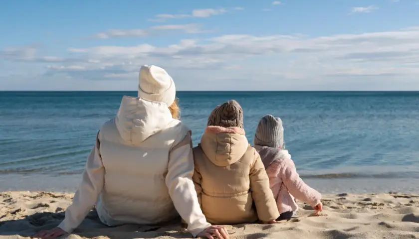 Rodzina w ciepłych ubraniach siedzi na plaży, patrząc na spokojne morze. Pogoda w czerwcu nad morzem bywa kapryśna, ale widok jest piękny.