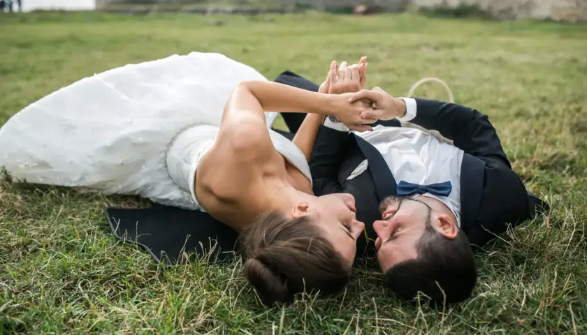 Para młoda leży na trawie podczas sesji plenerowej ślubnej, trzymając się za ręce. W tle ruiny zamku.
