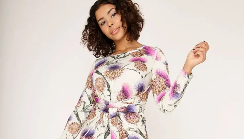 A woman with curly brown hair wears a long-sleeved white dress with a purple and pink thistle floral print.
