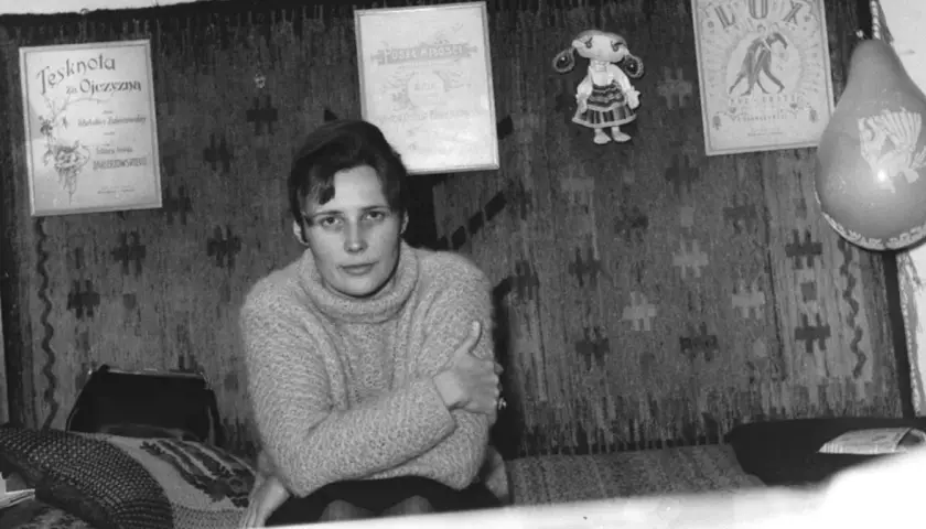 Młoda kobieta w swetrze siedzi na tle gobelinu. Na ścianie wiszą dyplomy i lalka. Czy to Agnieszka Osiecka, której prawdziwe nazwisko brzmi...