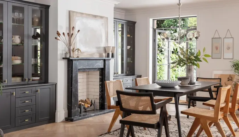 Modern dining room with dark grey cabinets, marble fireplace, and a dark wood table with cane-backed chairs, showcasing current trends in dining room furniture.
