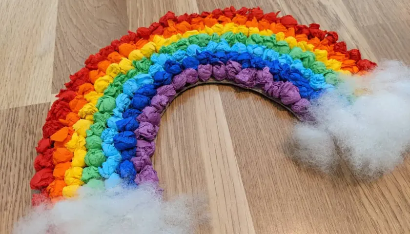 Ein bunter Regenbogen, gebastelt aus zerknülltem Krepppapier in allen Farben, mit flauschigen Wolken aus Watte.