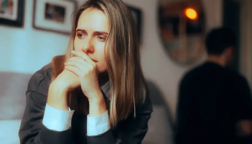 Smutna kobieta z rękami przy twarzy, siedząca z dala od mężczyzny. Czuć dystans po kłótni.