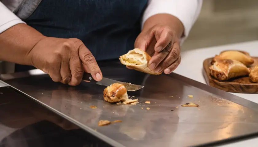 Manos cortan una empanada dorada, revelando su relleno. Otra masa empanada espera en una tabla.