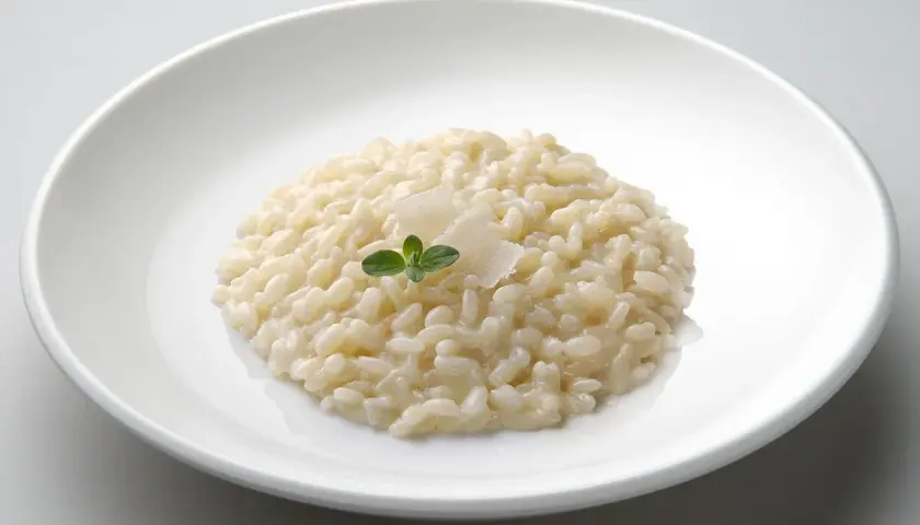 Un cremoso risotto in cagnone, guarnito con parmigiano e una fogliolina verde, servito in un piatto bianco.