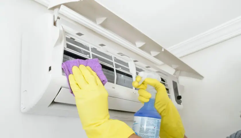 Man con guantes amarillos limpia un aire acondicionado con un paño morado y spray. Aprende como desatascar un aire acondicionado.