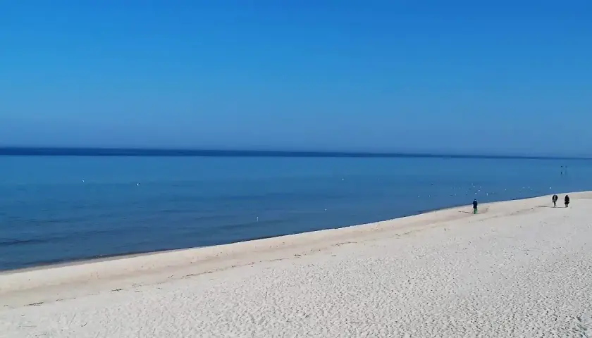 Grzybowo nadmorskie: zejście na plażę. Dwie osoby spacerują po piaszczystej plaży, morze i błękitne niebo.