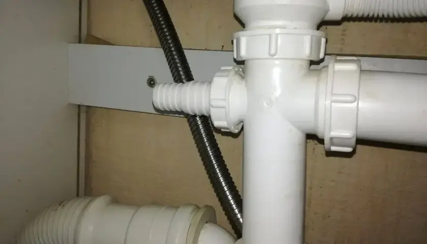 Biały plastikowy trójnik hydrauliczny pod zlewem, do którego podłączona jest elastyczna rura odpływowa i metalowy wąż.