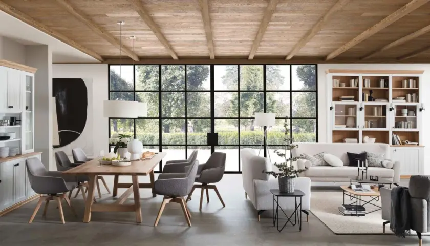 Geräumiges Esszimmer mit Holzbalkendecke im Bauernhaus Stil, Holztisch, grauen Stühlen und großer Fensterfront mit Blick ins Grüne.