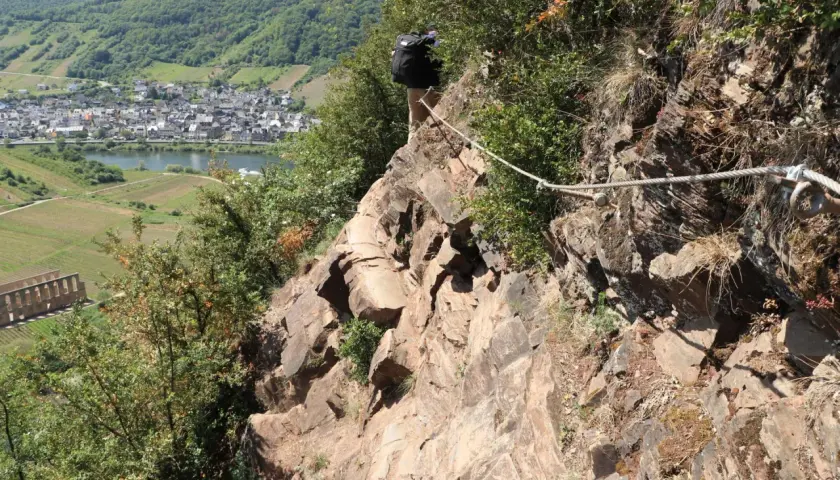Wanderer sichert sich mit Seil am Felssteig über der Mosel. Blick auf Weinberge und Dorf.