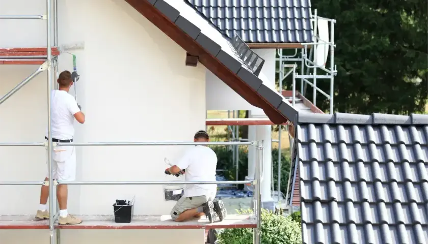 Dwóch mężczyzn na rusztowaniu maluje białą elewację. Sprawdzają tynk silikonowy cena za m2 robocizny.