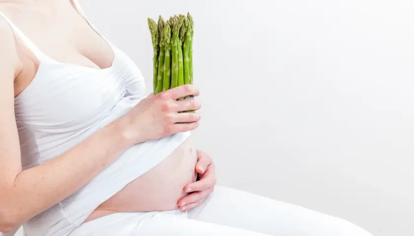 Ciężarna kobieta w białej sukience trzyma pęczek zielonych szparagów, symbol zdrowej diety w ciąży.