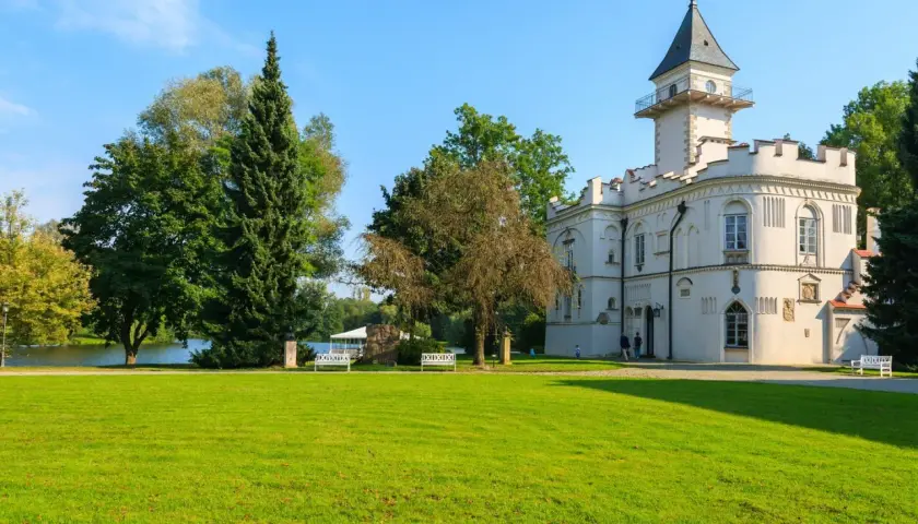 Biały zamek z wieżą, otoczony zielonym parkiem i jeziorem. Urokliwe zamki na Mazowszu zachwycają.
