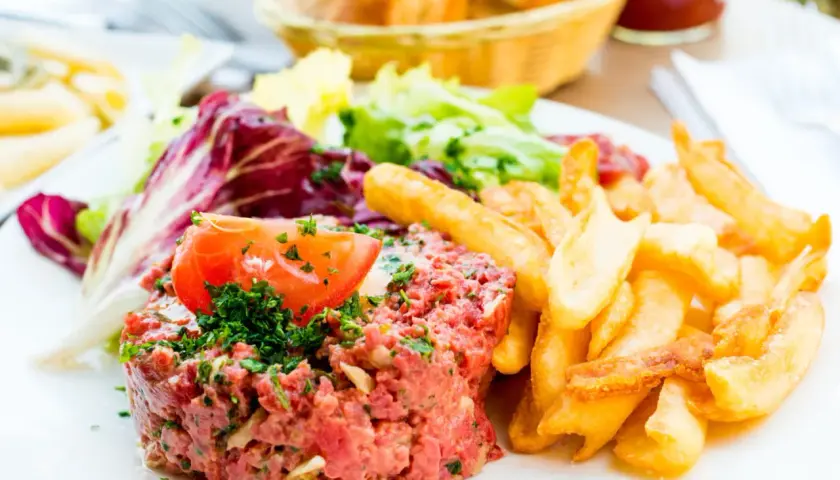 Un délicieux tartare de boeuf, garni d'une tranche de tomate et de persil, servi avec des frites croustillantes et une salade fraîche.