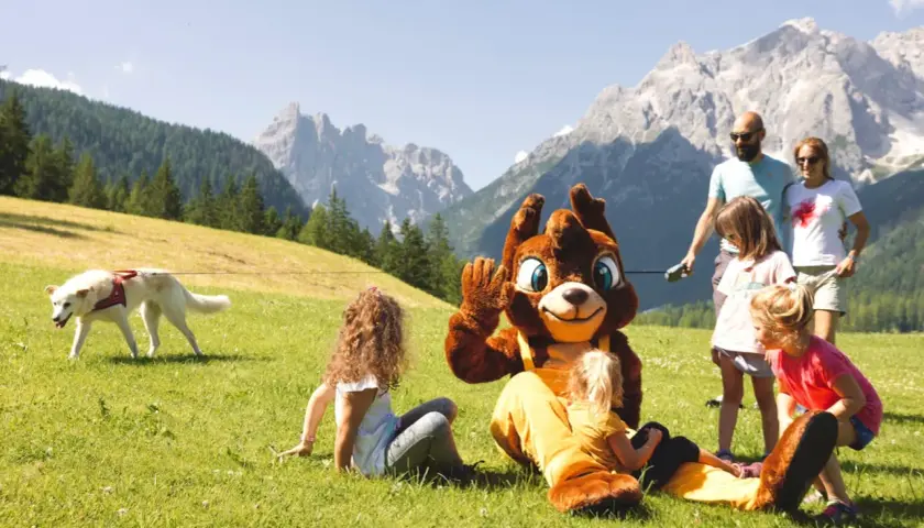 Famiglia al family resort Rainer Sesto, con mascotte e cane su prato verde, montagne sullo sfondo.
