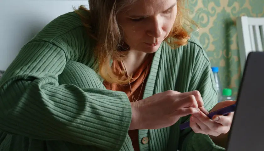Kobieta w zielonym swetrze przegląda dokumenty na telefonie, być może wniosek o zasiłek chorobowy.