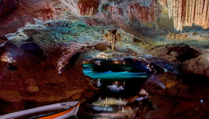 Interior de cueva con estalactitas, agua turquesa y una barca. Consulta las cuevas de Vall d'Uixó horarios y precios para tu visita.