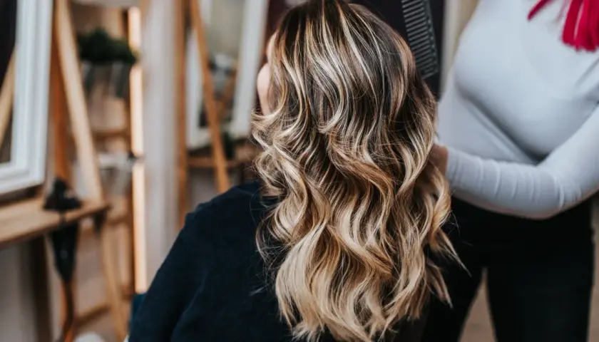 Mujer con cabello castaño y mechas rubias ondulado, visto desde atrás en un salón de belleza.