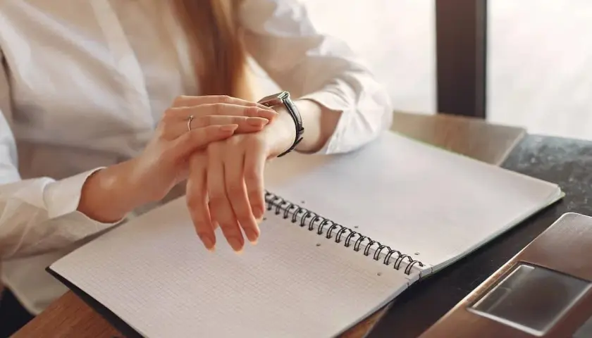 Kobieta w białej koszuli sprawdza zegarek, pracując po godzinach. Na biurku leży otwarty notatnik i laptop.