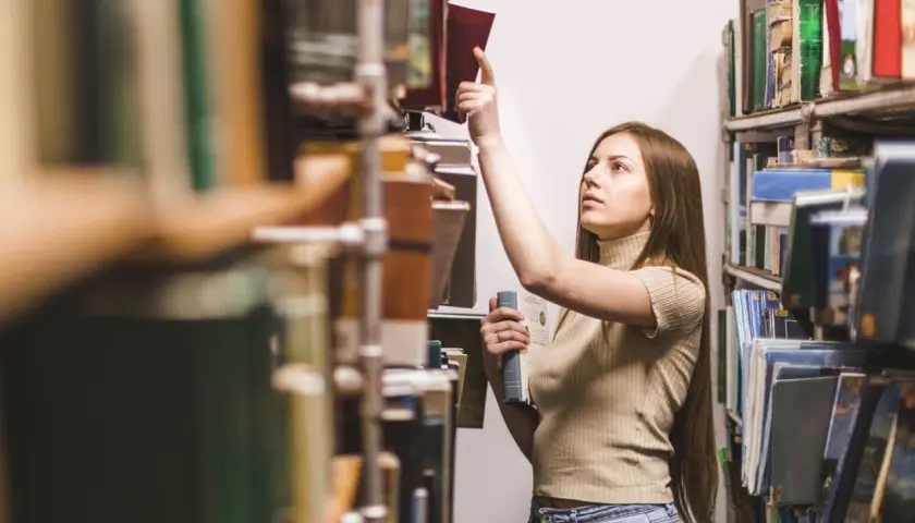 Młoda kobieta w bibliotece szuka inspiracji, zastanawiając się, jakie gatunki literackie są najbardziej poszukiwane przez wydawców w 2026.