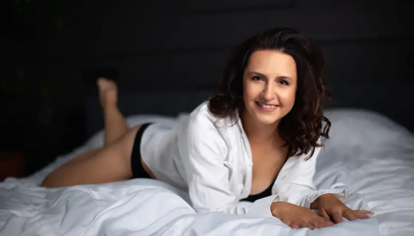Uśmiechnięta kobieta w białej koszuli leży na łóżku. Świetne pomysły na sesję w domu.