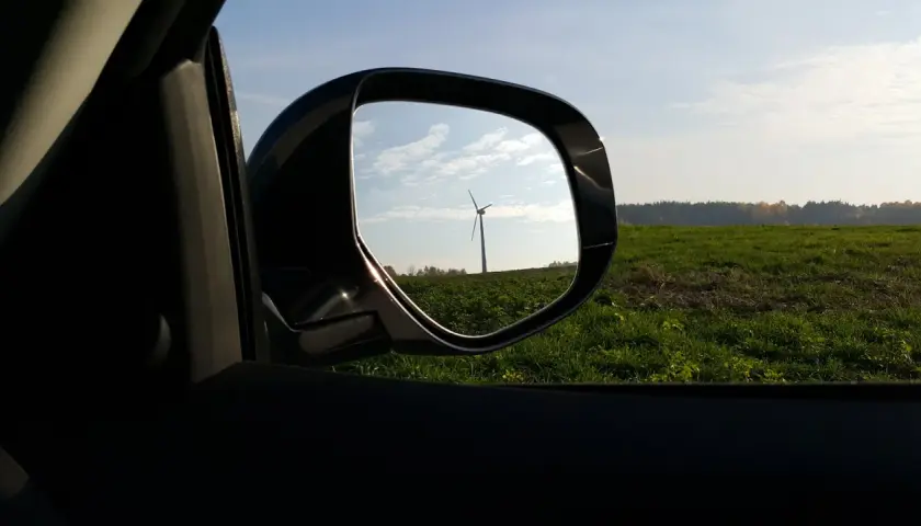 Widok w lusterku samochodowym na wiatrak na tle zielonego pola i nieba.
