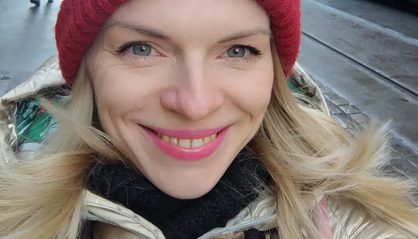Uśmiechnięta blondynka w czerwonej czapce i złotej kurtce, jakby właśnie spacerowała po ulicach Czech, gdzie ludność cieszy się zimową aurą.