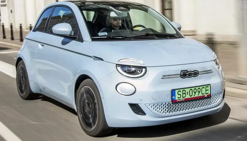 Jasnoniebieski fiat elektryczny 500 z czarnym dachem jedzie ulicą. Kierowca w okularach przeciwsłonecznych prowadzi auto.