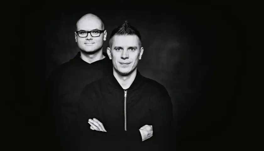 Dwóch mężczyzn w czarnych bluzach na ciemnym tle. Jeden z nich ma okulary, drugi irokeza. Wyglądają jakby właśnie wylądowali po locie dzika.