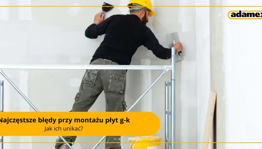 Budowlaniec w żółtym kasku i roboczych spodniach na rusztowaniu wygładza ścianę gładzią. Unikaj błędów przy montażu płyt g-k, jak przy nitowaniu.