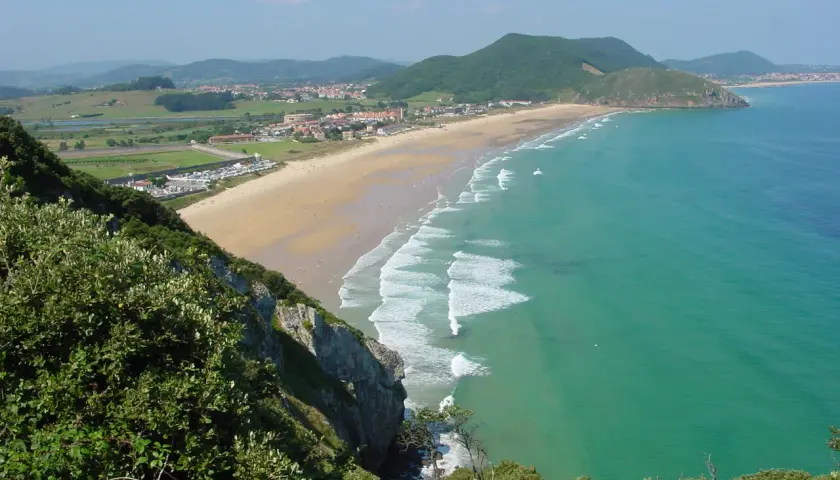 Vista panorámica de la playa de Berria, con arena dorada, aguas turquesas y colinas verdes al fondo.