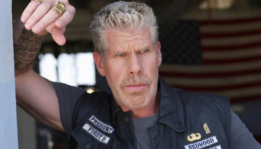 Ron Perlman w skórzanej kamizelce z naszywkami, z tatuażem na ramieniu i pierścieniami na palcach, na tle amerykańskiej flagi.