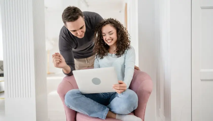 Para przygotowuje się do ślubu, korzystając z nauk przedmałżeńskich online. Uśmiechnięci patrzą na ekran laptopa.