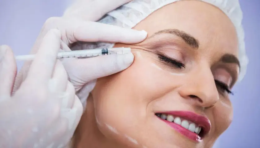 Kobieta uśmiecha się podczas zabiegu baby botox. Dłoń w rękawiczce trzyma strzykawkę z igłą, aplikując preparat w okolicę oka.