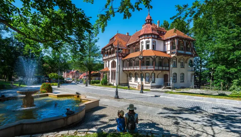 Dwie osoby siedzą przy fontannie, podziwiając architekturę luksusowego sanatorium w otoczeniu zieleni.