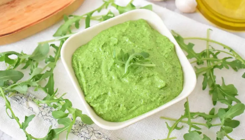 Aksamitne, zielone pesto z rukoli w białej miseczce, otoczone świeżymi listkami rukoli, czosnkiem i oliwą.