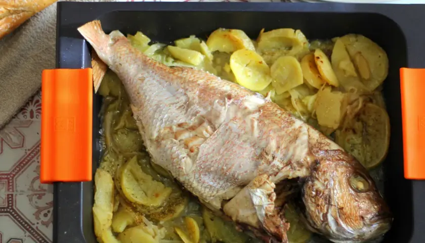 Pargo entero al horno con patatas y cebolla en bandeja negra con asas naranjas.