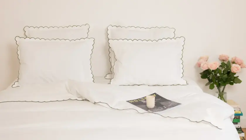 Parure de lit blanche avec passepoil vert, idéale pour un lit 140x190. Un magazine et des roses apportent une touche décorative.
