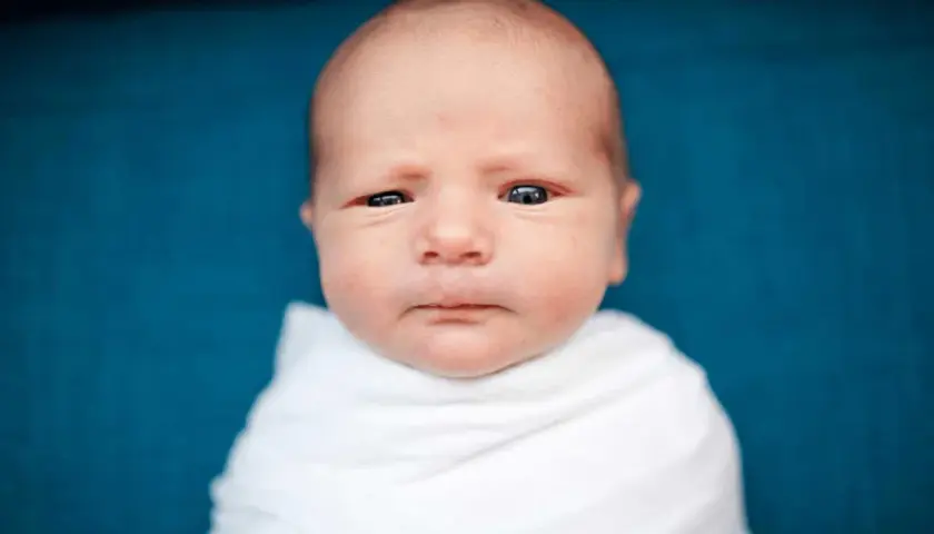 Un neonato senza capelli davanti, avvolto in una coperta bianca, guarda con occhi curiosi.